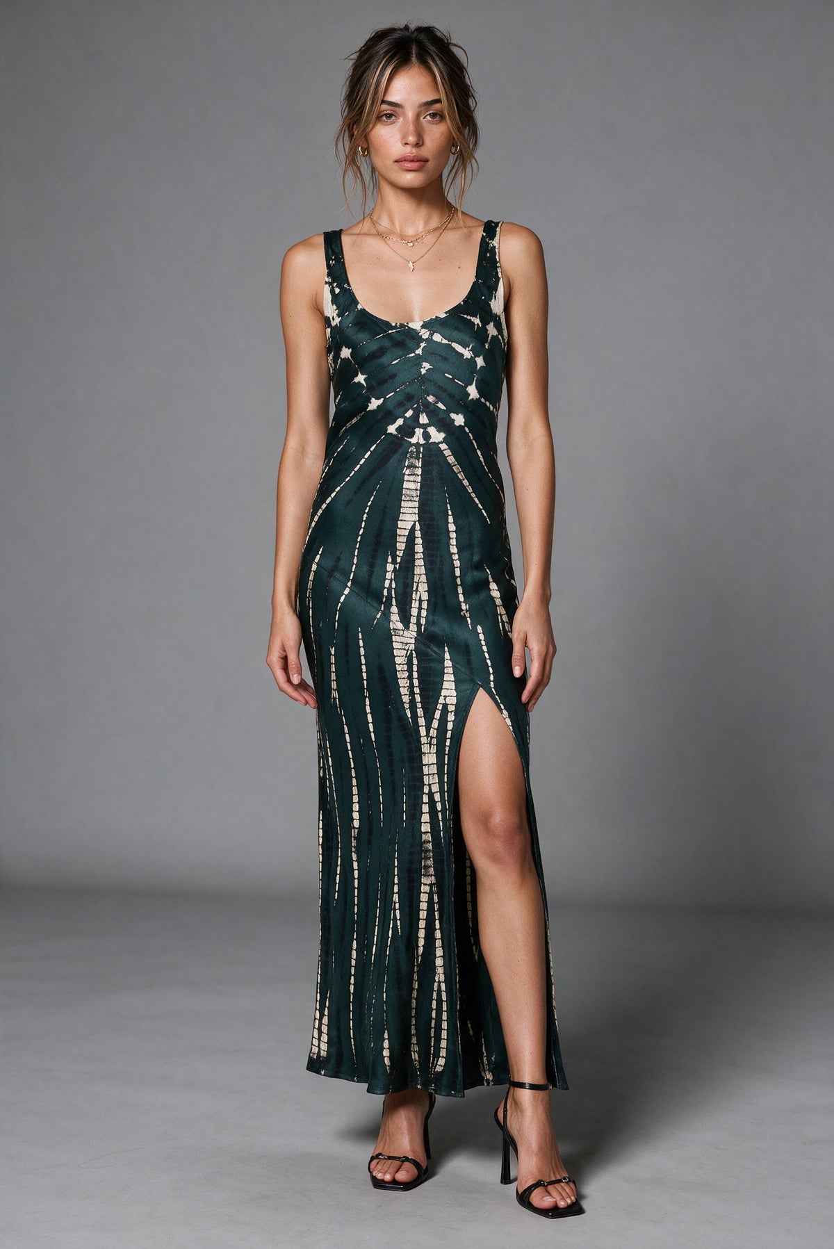 A woman modeling a sleeveless dark green midi dress with a vertical cream tie-dye pattern and a high side slit, standing against a neutral gray studio background.