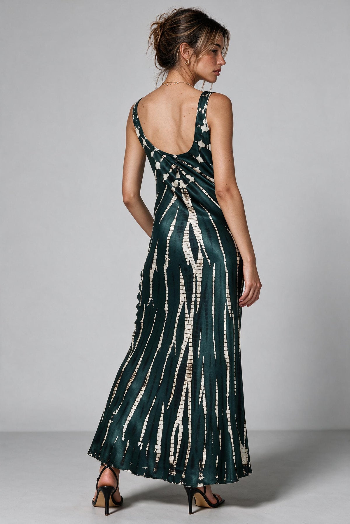 A woman modeling a sleeveless midi dress from the back, featuring a dark green base with vertical white tie-dye stripes and a wrap-style back, set against a plain gray studio background.