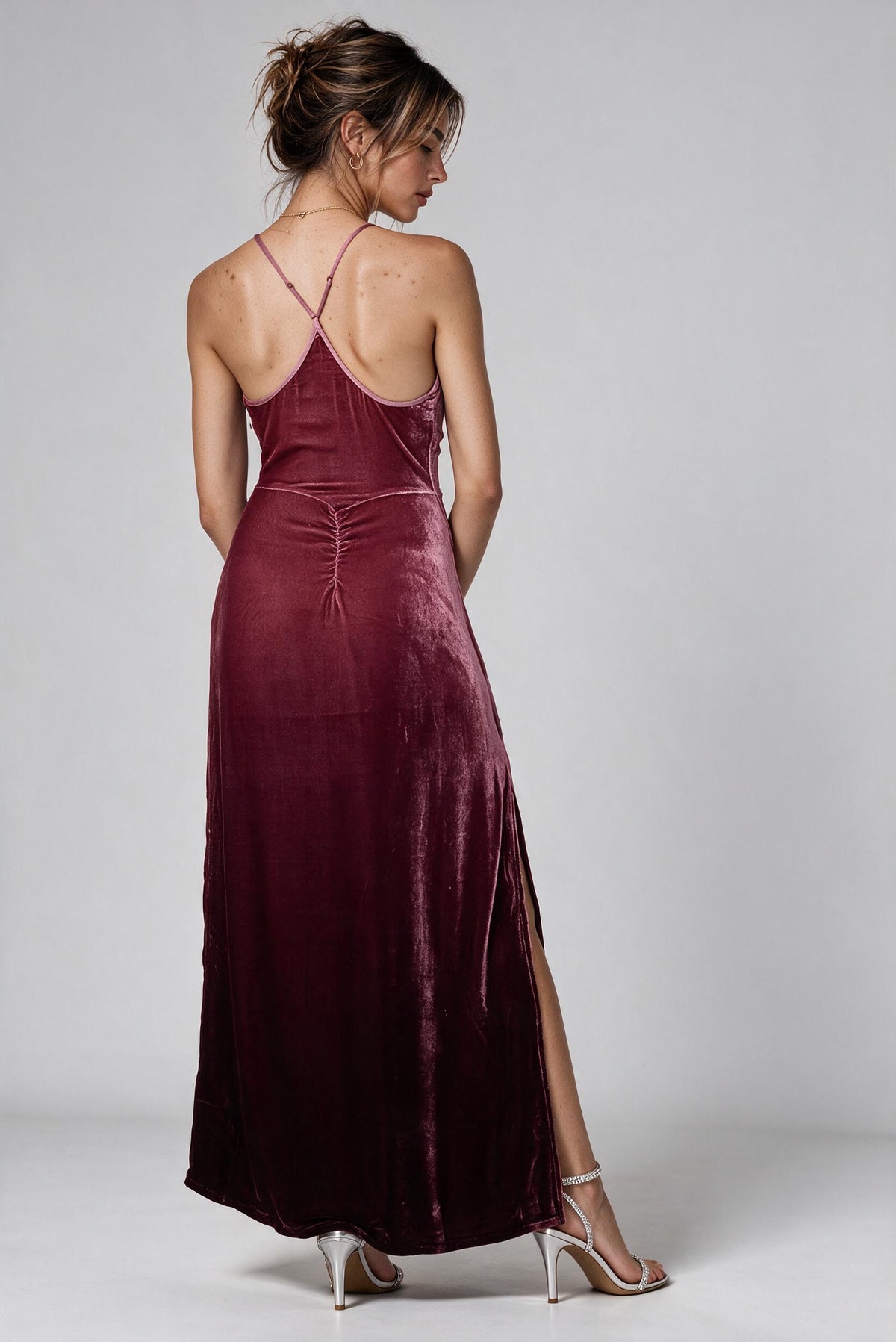 A woman modeling a floor-length burgundy velvet maxi dress from the back, featuring thin double straps, a gathered waist, and a high side slit, paired with clear high-heeled sandals against a light gray studio background.