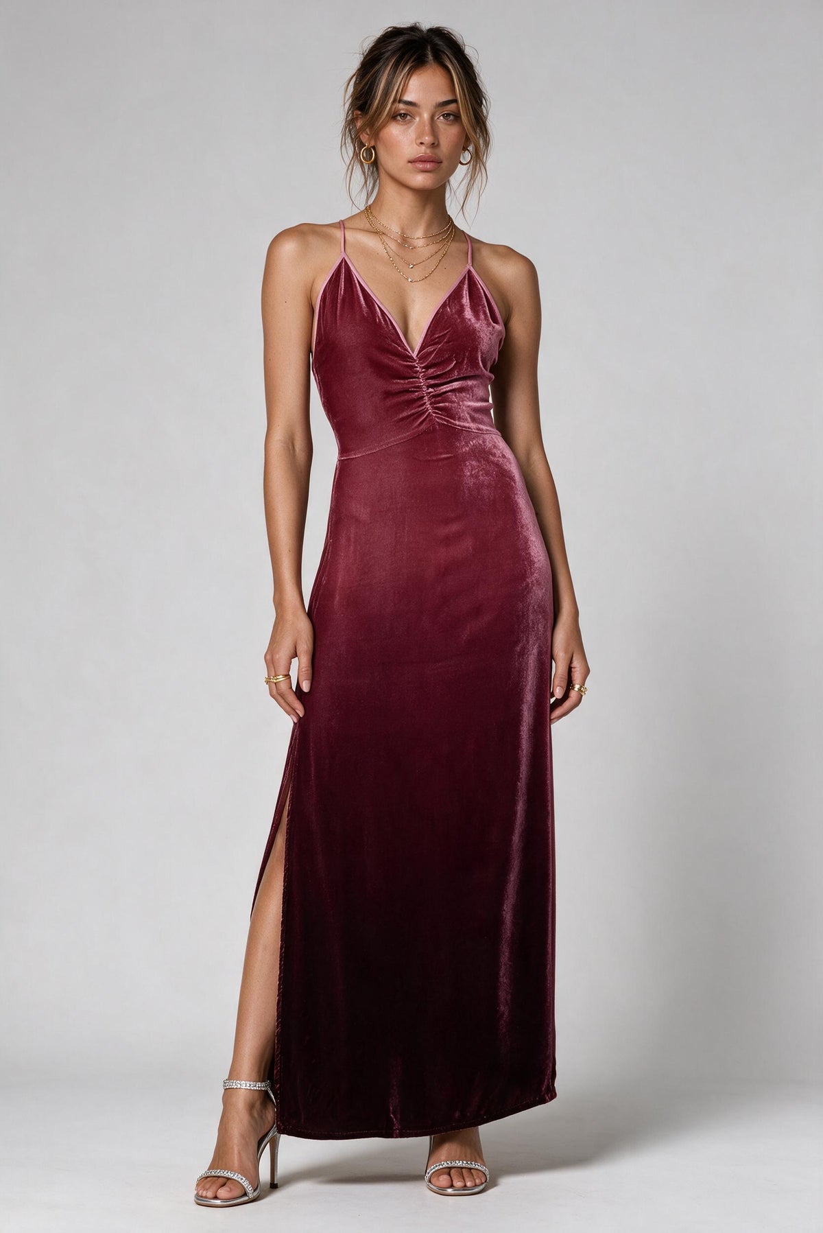 A woman modeling a floor-length burgundy velvet slip dress with thin straps, a gathered sweetheart neckline, and a thigh-high slit, photographed against a light gray studio background.