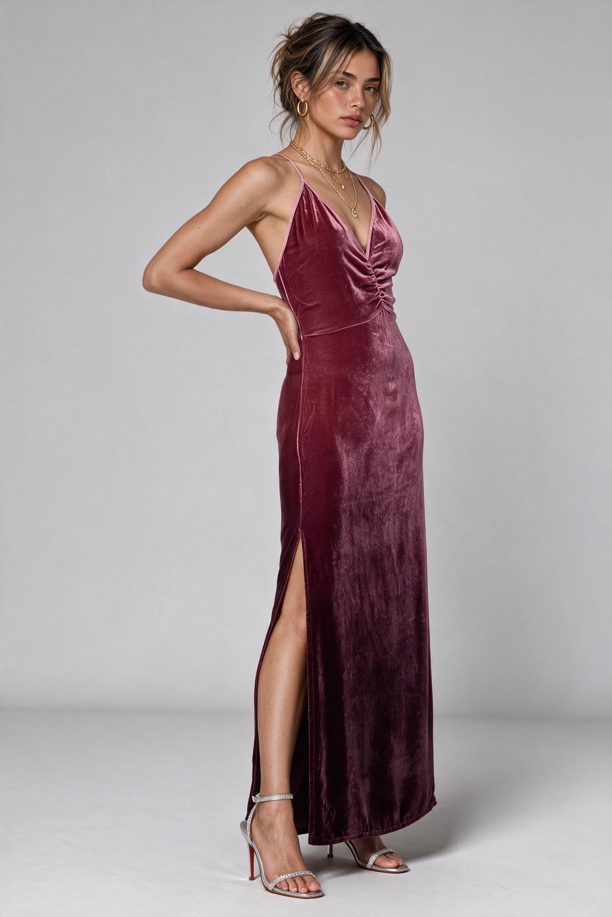 A woman modeling a floor-length plum velvet halter gown with a ruched bodice, thigh-high slit, and silver strappy heels against a light gray studio background.