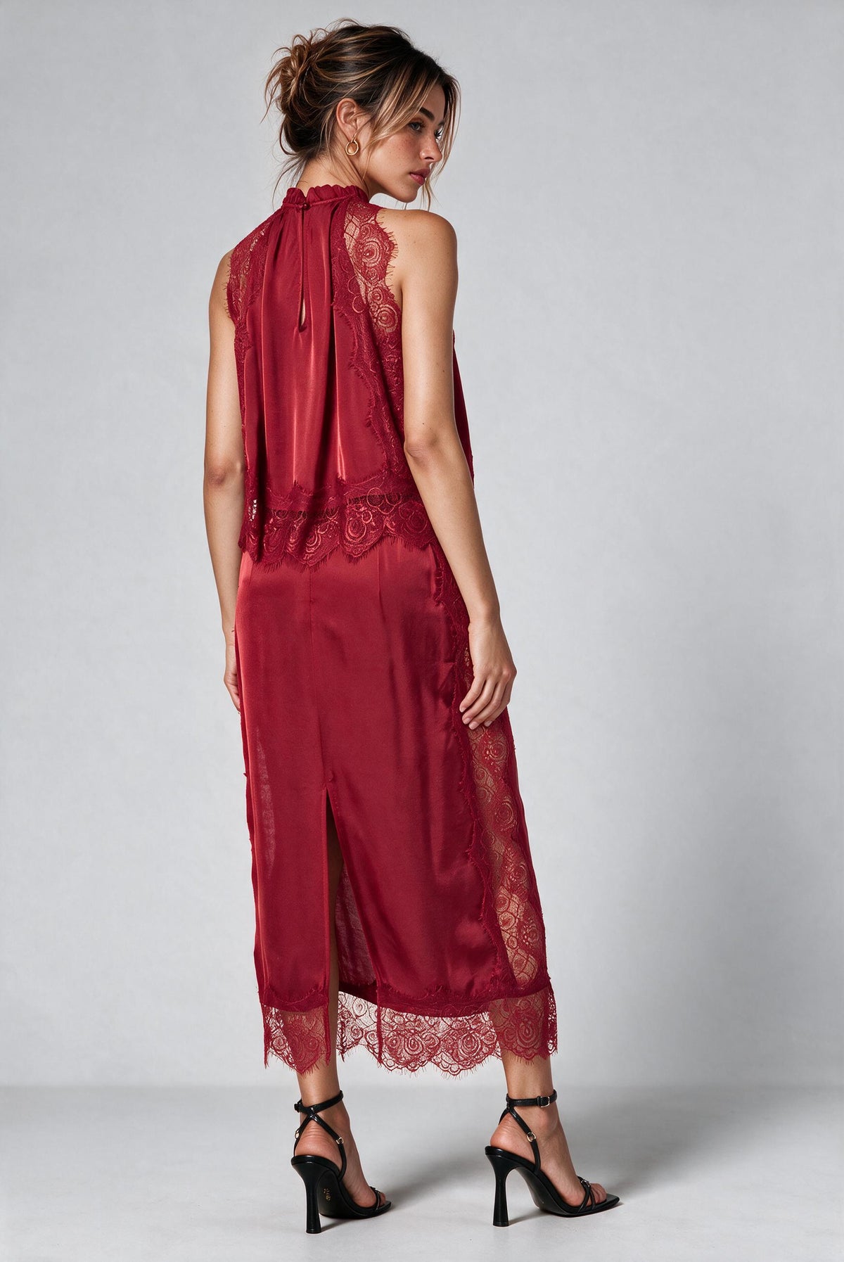 A woman viewed from the backside modeling a sleeveless high-neck top and matching skirt in deep burgundy satin with lace trim and back slit, paired with black strappy high-heeled sandals against a light gray studio background., 