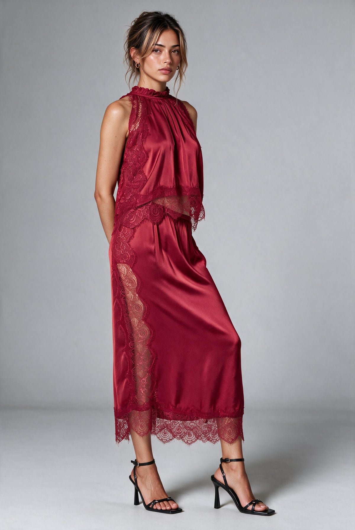 A woman modeling a sleeveless high-neck top and matching skirt in deep burgundy satin with lace trim and back slit, paired with black strappy high-heeled sandals against a light gray studio background., 