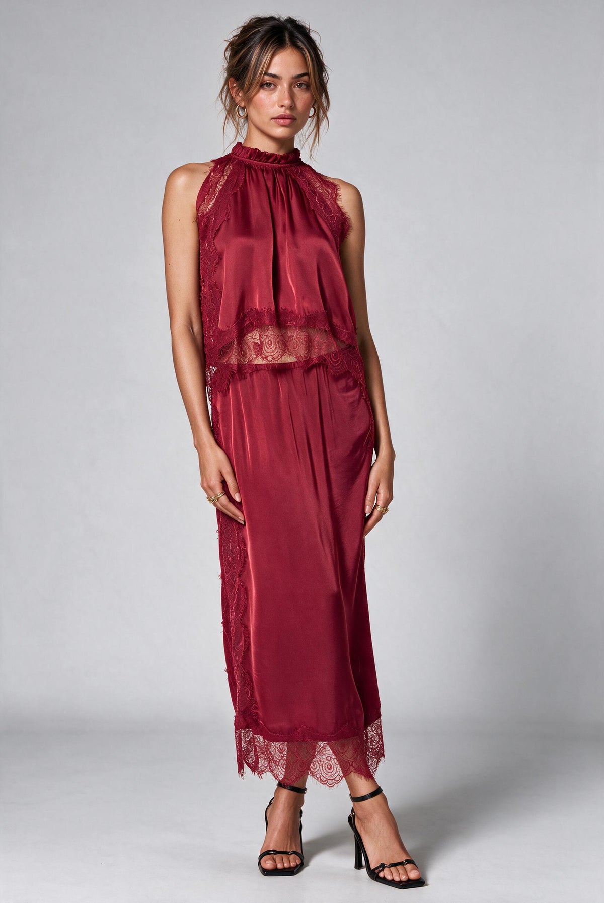 A woman modeling a sleeveless high-neck top and matching skirt in deep burgundy satin with lace trim and back slit, paired with black strappy high-heeled sandals against a light gray studio background., 