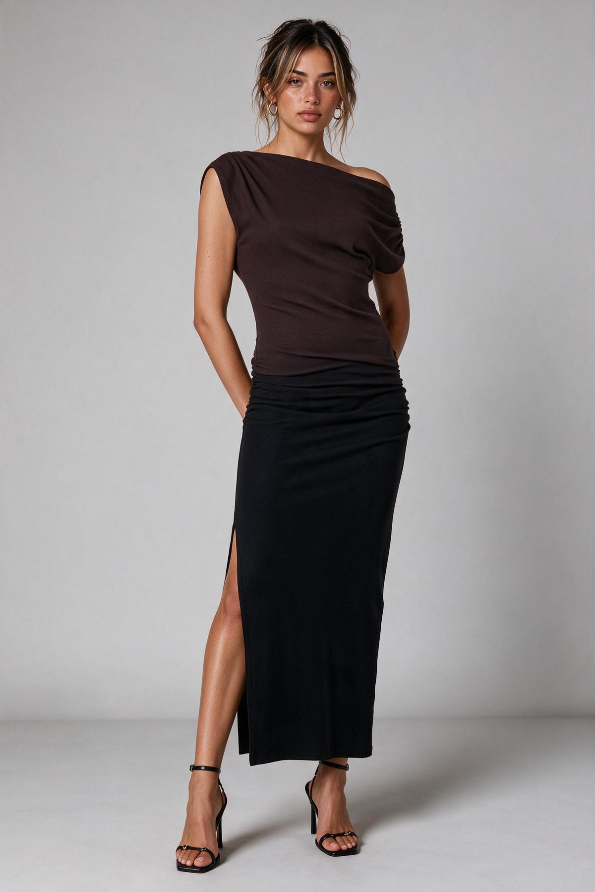 A woman modeling a dark brown ombre off-the-shoulder top paired with a long black ruched skirt featuring a thigh-high slit, styled with black strappy high-heeled sandals against a neutral gray studio background.