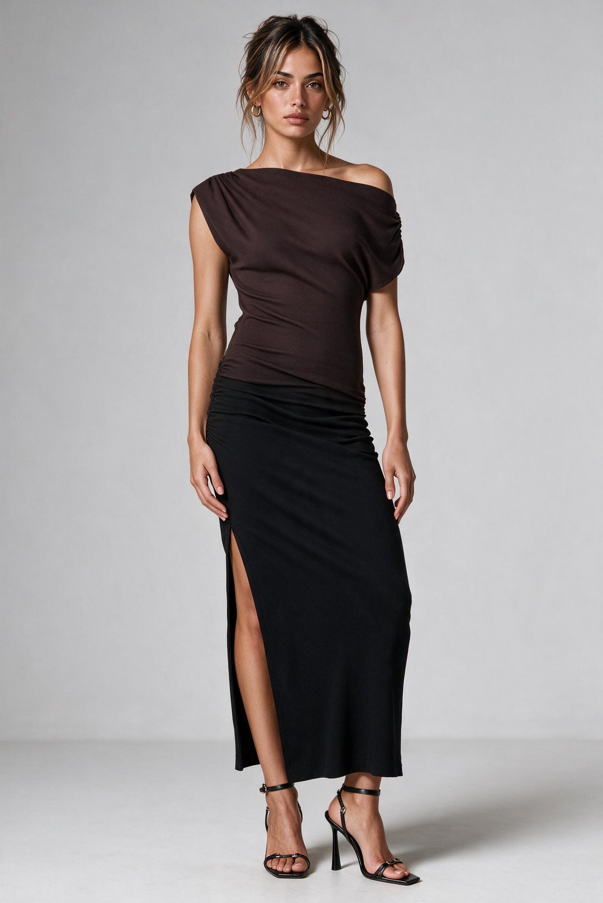 A woman modeling a dark brown ombre off-the-shoulder dress  featuring a thigh-high slit, styled with black strappy heels against a light gray studio background.