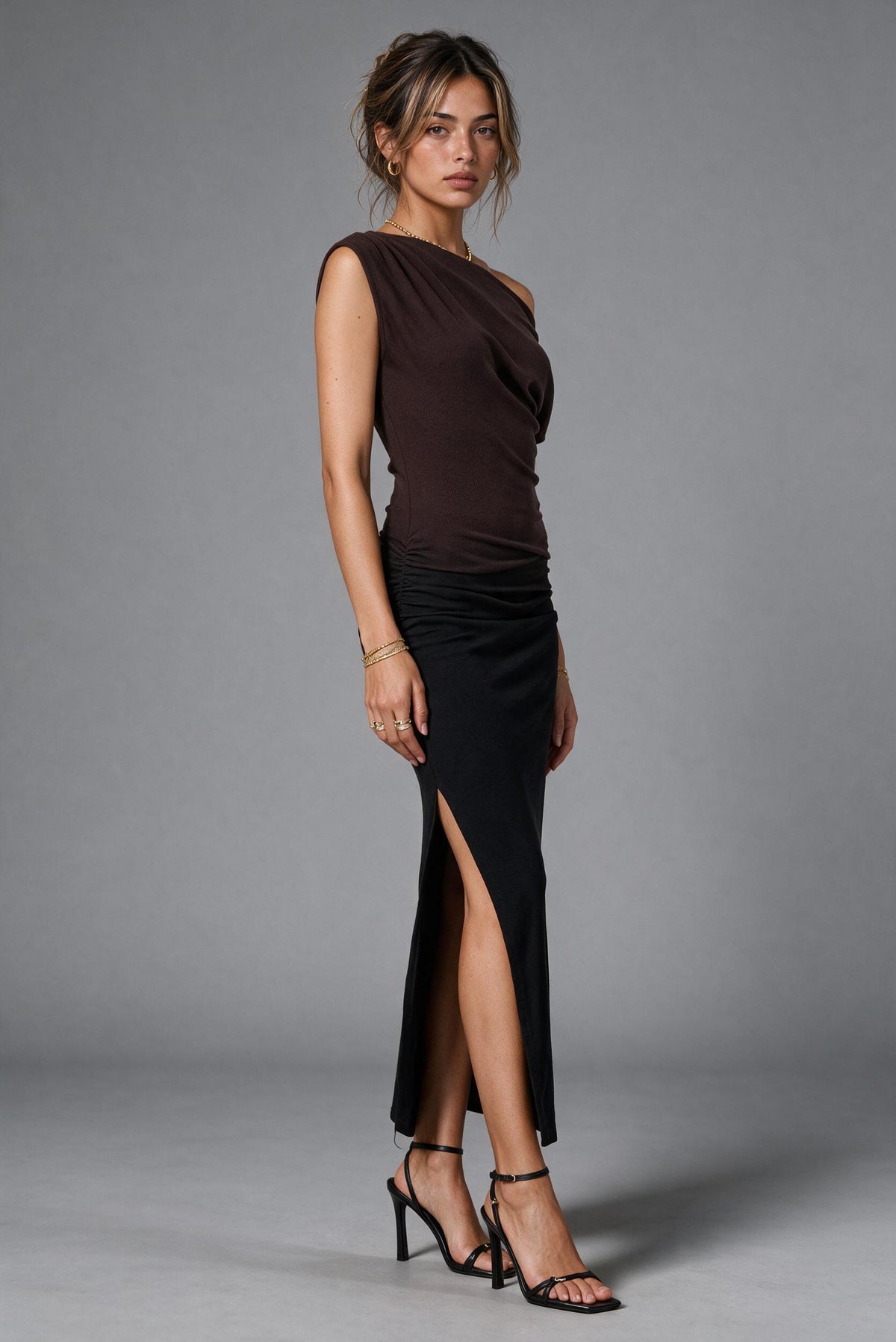 A woman modeling a dark brown ombre one-shoulder draped dress with a black high-slit skirt, paired with black strappy heels, against a solid gray studio background.