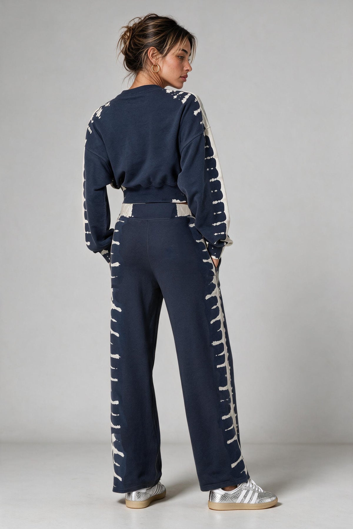 Backside of a model standing against a light gray studio background wearing a matching navy blue and off-white tie-dye two-piece lounge set consisting of a cropped long-sleeve top and wide-leg pants, paired with metallic silver sneakers.