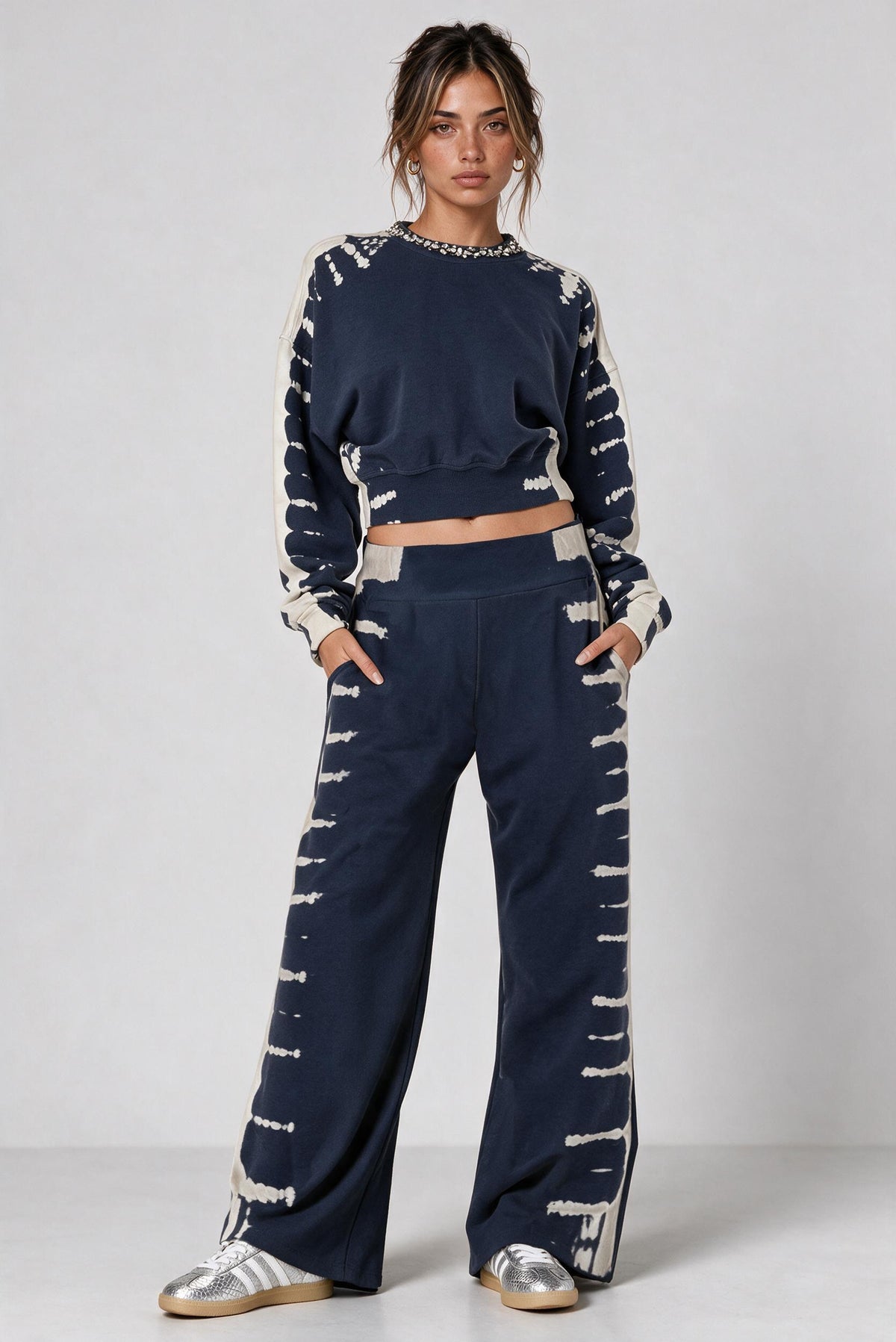 A model stands against a light gray studio background wearing a matching navy blue and off-white tie-dye two-piece lounge set consisting of a cropped long-sleeve top and wide-leg pants, paired with metallic silver sneakers.