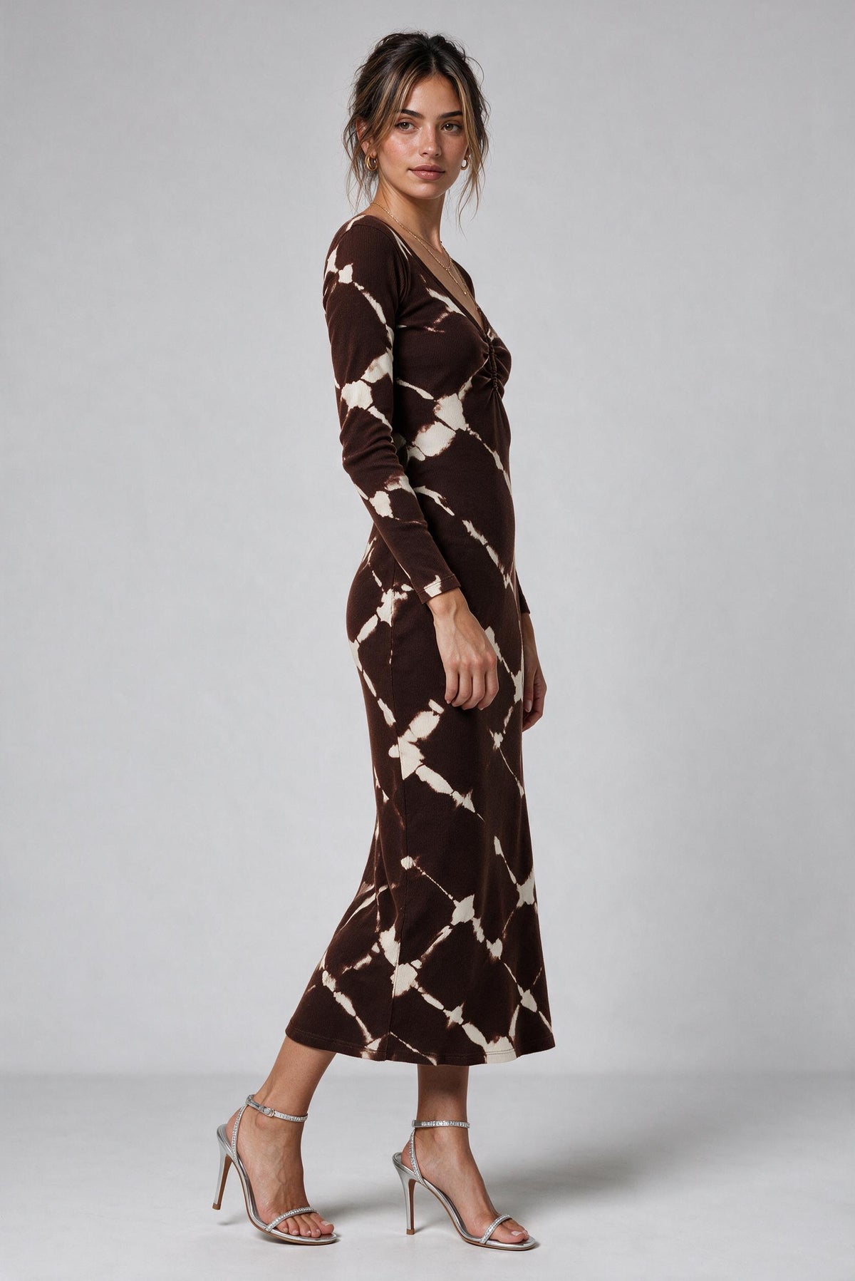 A woman modeling a long-sleeve midi dress with a brown and cream tie-dye pattern, paired with silver high-heeled sandals, against a light gray studio background.