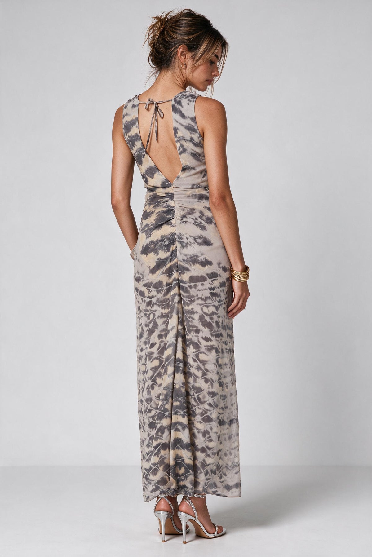 A woman shown from the back wearing a sleeveless maxi dress with a tie-shoulder open back and an abstract animal-like print in beige, gray, and brown tones, photographed against a light gray studio background.