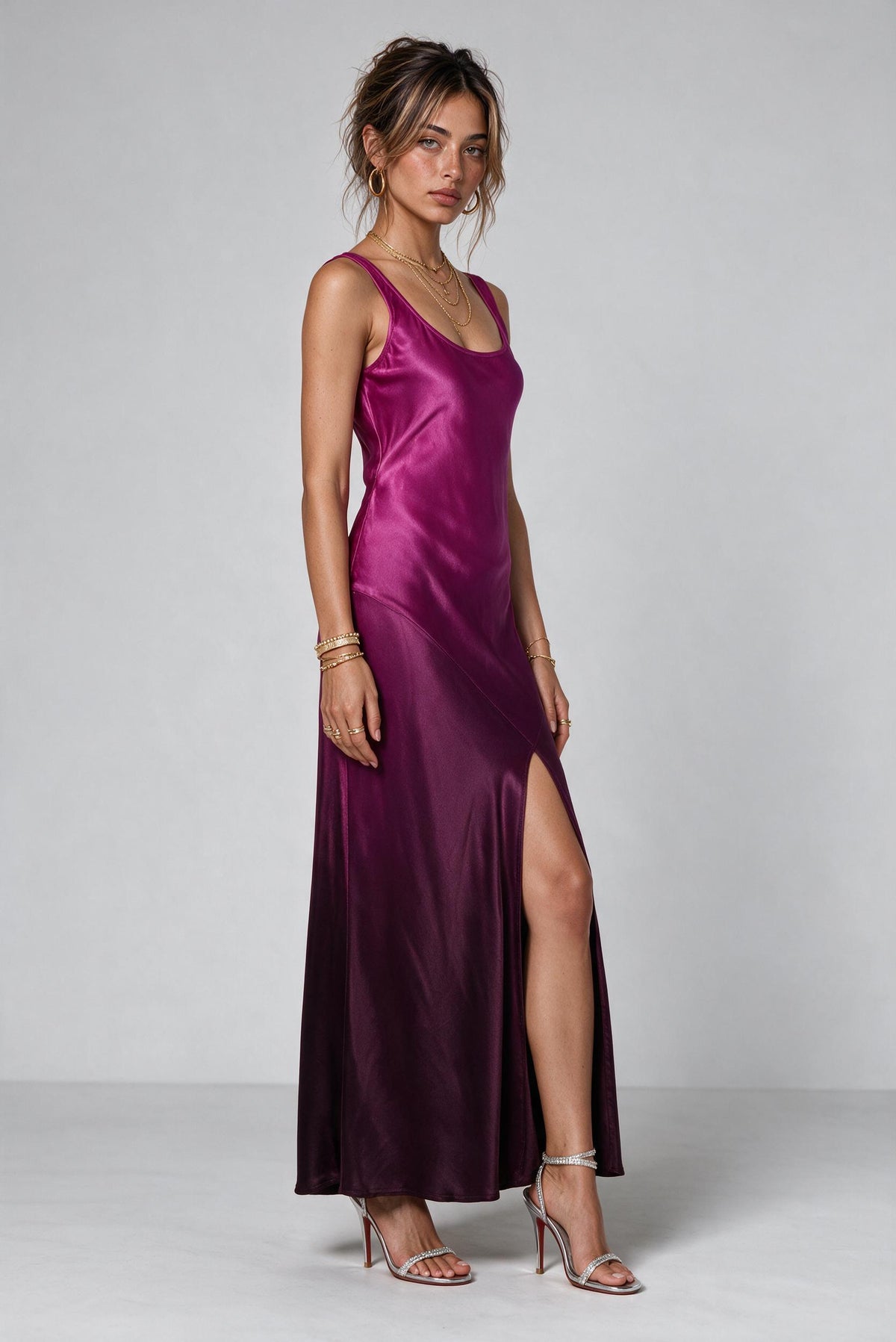 A woman modeling a floor-length satin slip dress in a rich magenta-purple color, shown from the back against a neutral gray studio background.