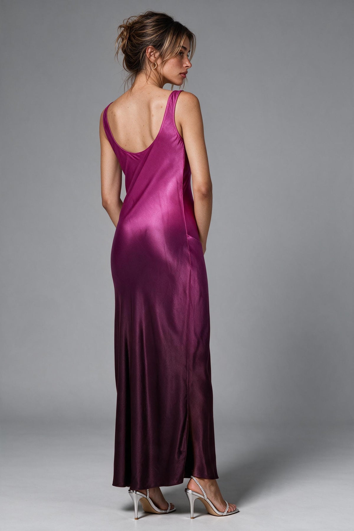 A woman modeling a floor-length satin slip dress in a rich magenta-purple color, shown from the back against a neutral gray studio background.