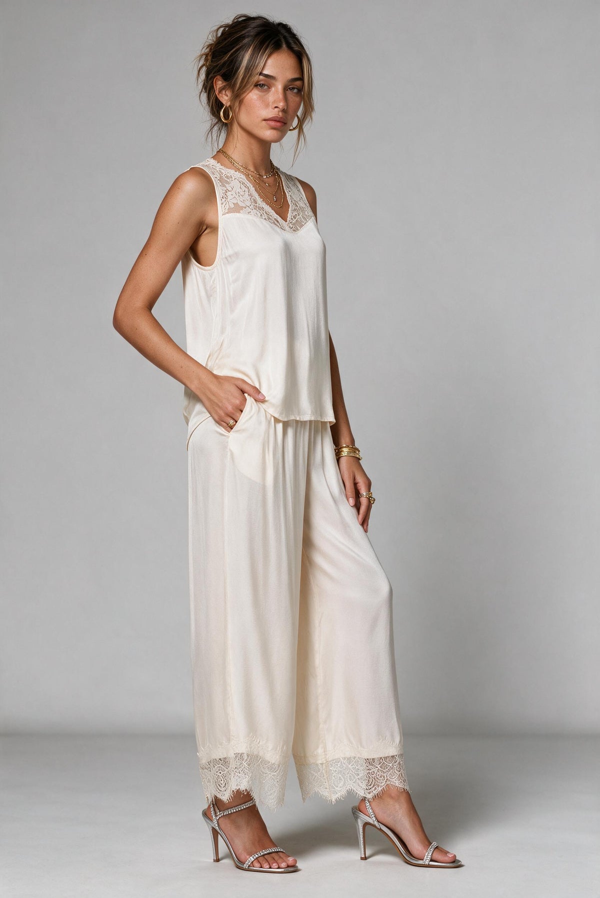A woman models a two-piece cream satin pajama set featuring a sleeveless top with lace trim and matching wide-leg pants with lace hems, paired with silver high-heeled sandals against a light gray studio background.