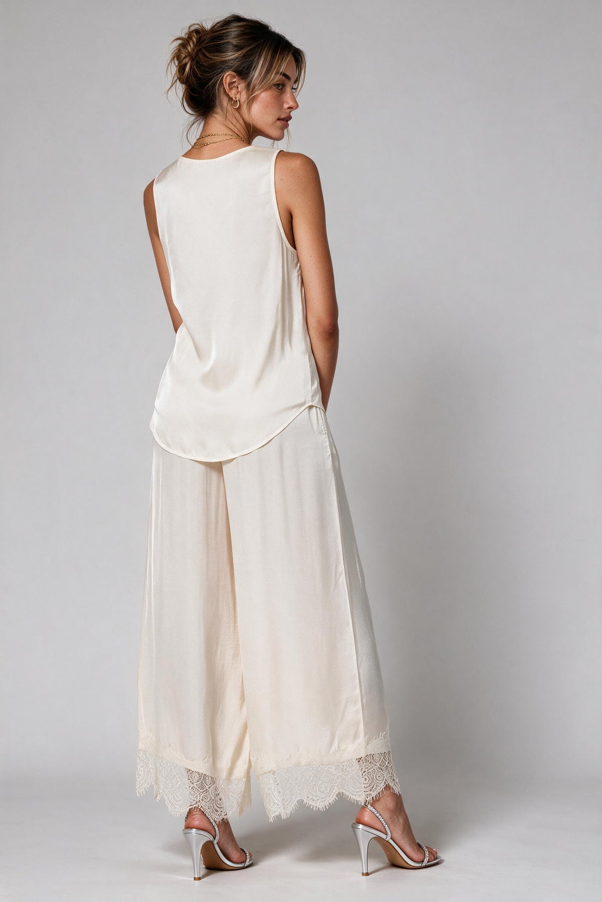 A woman shown from the back wearing a sleeveless cream top and matching wide-leg pants with lace trim, paired with clear high-heeled sandals, against a light gray studio background.