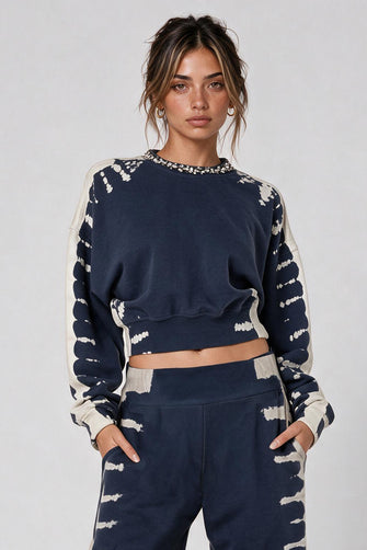 A model stands against a light gray studio background wearing a matching navy blue and off-white tie-dye two-piece lounge set consisting of a cropped long-sleeve top and wide-leg pants, paired with metallic silver sneakers.
