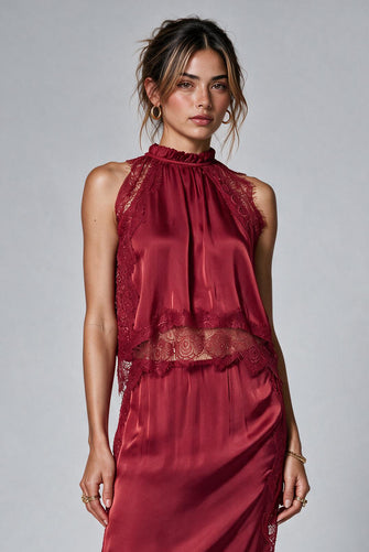 A woman modeling a sleeveless high-neck top and matching skirt in deep burgundy satin with lace trim.  Paired with black strappy high-heeled sandals against a light gray studio background.