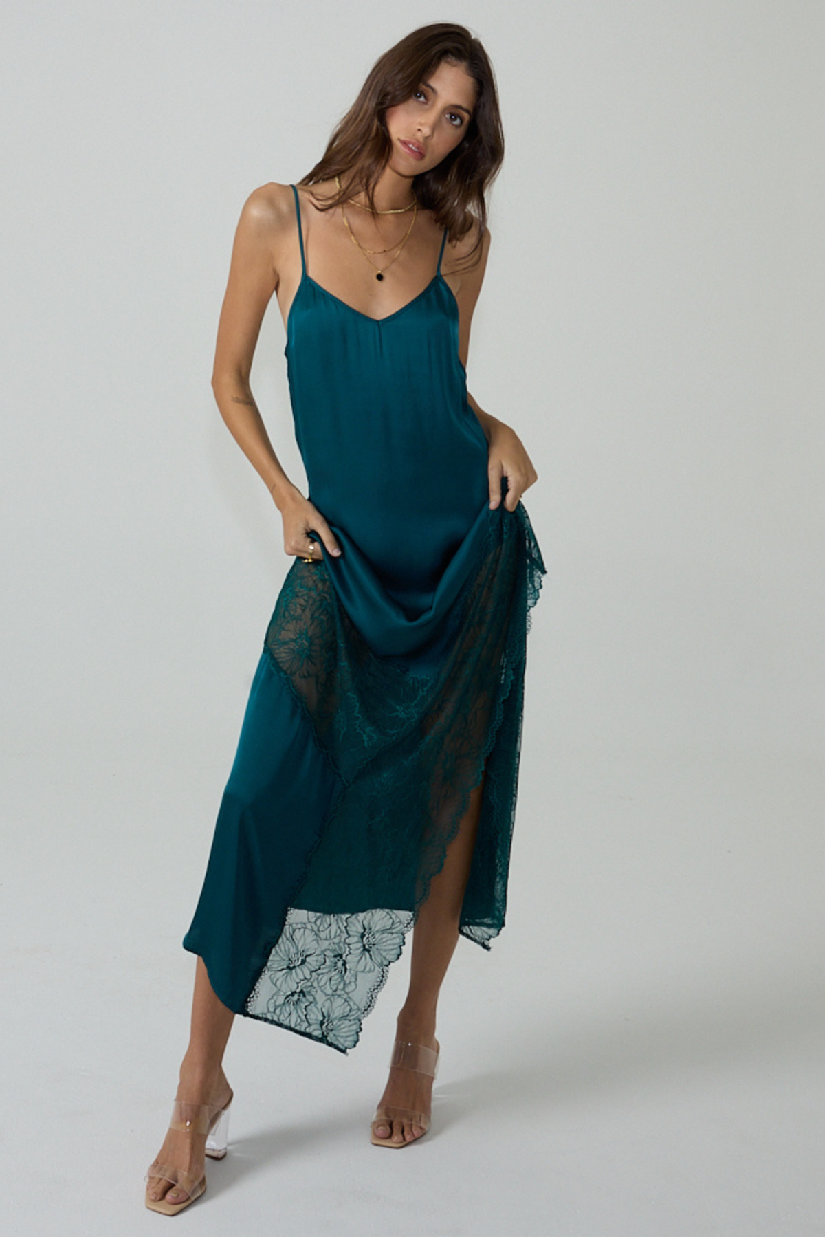A woman modeling a teal green spaghetti-strap slip dress with a sheer lace overlay and a high-low hemline, set against a plain light gray studio background.