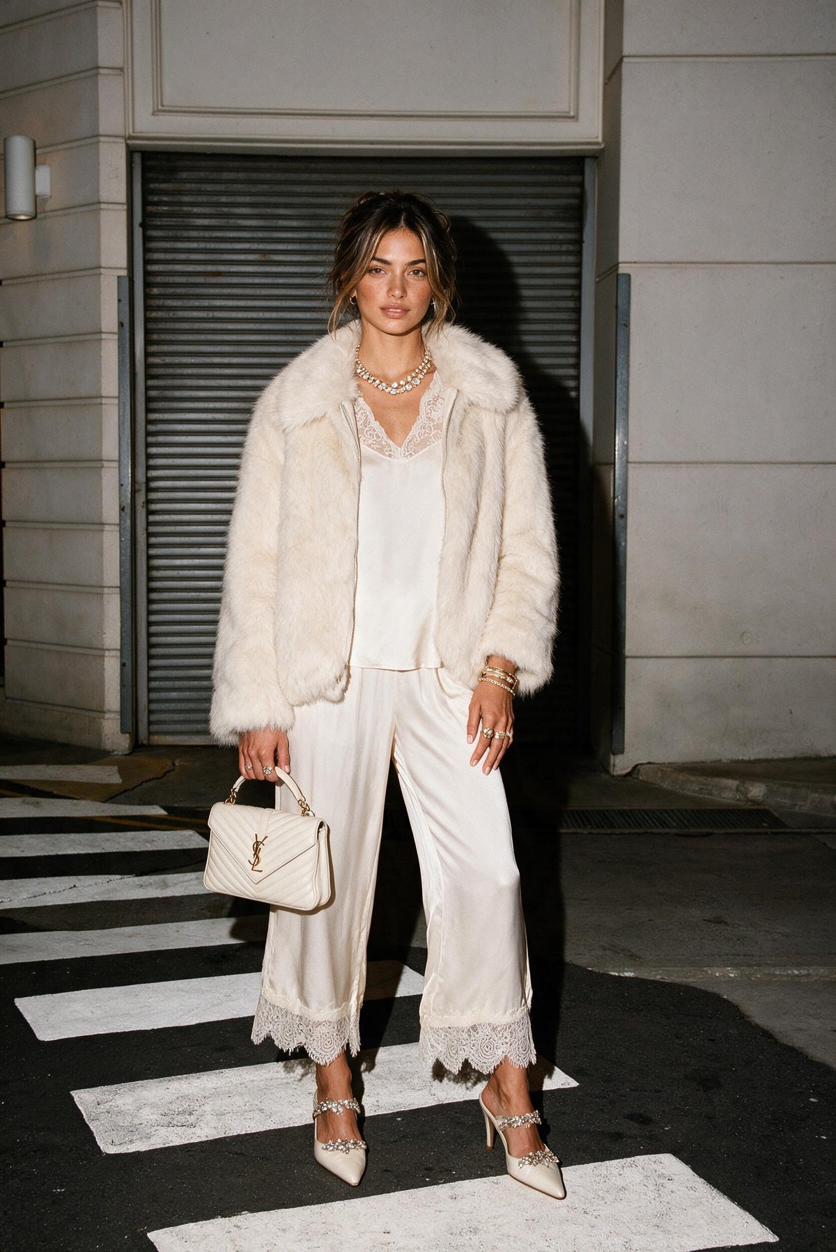 A woman stands on a city crosswalk at night wearing an off-white faux fur jacket over a matching cream satin outfit with lace-trimmed wide-leg pants and pointed heels. She carries a small structured cream handbag.