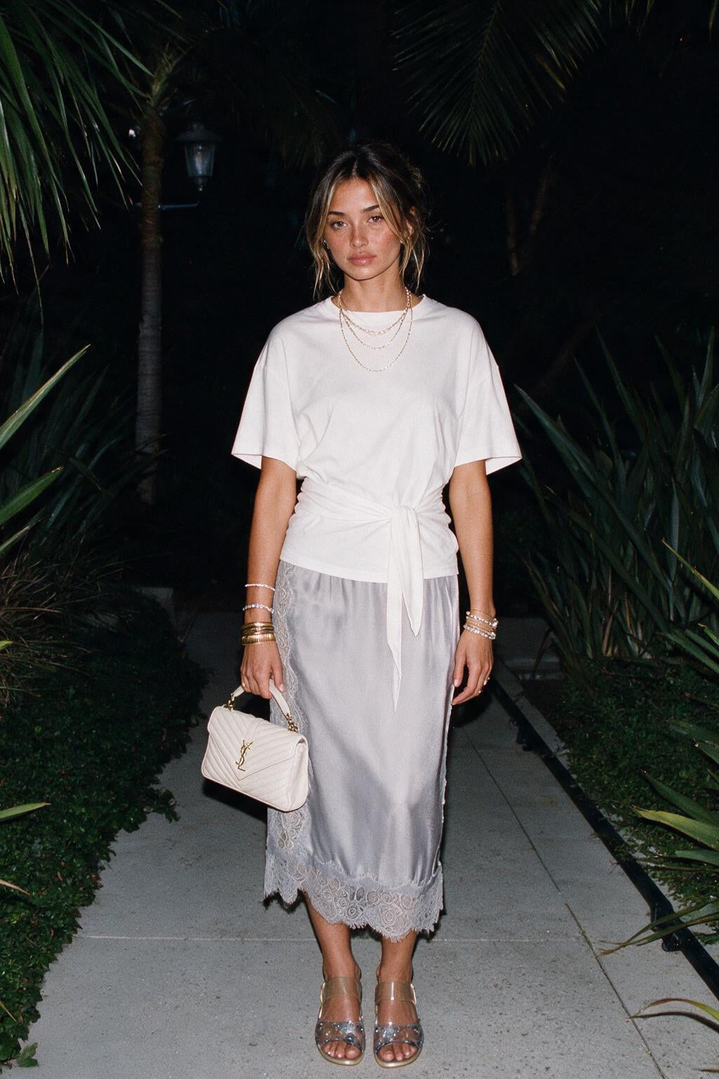 A woman stands on a nighttime outdoor walkway wearing a white short-sleeve wrap-style top and a sheer light gray midi skirt with lace trim. She is carrying a small white quilted handbag and wearing layered necklaces and bracelets.
