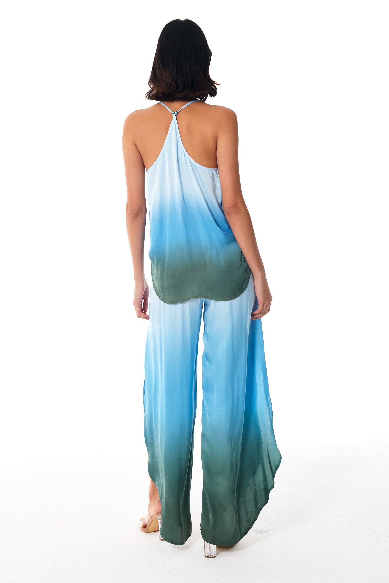 A woman viewed from the back wearing a sleeveless halter-style jumpsuit with a blue-to-green ombre gradient, photographed against a plain white studio background.