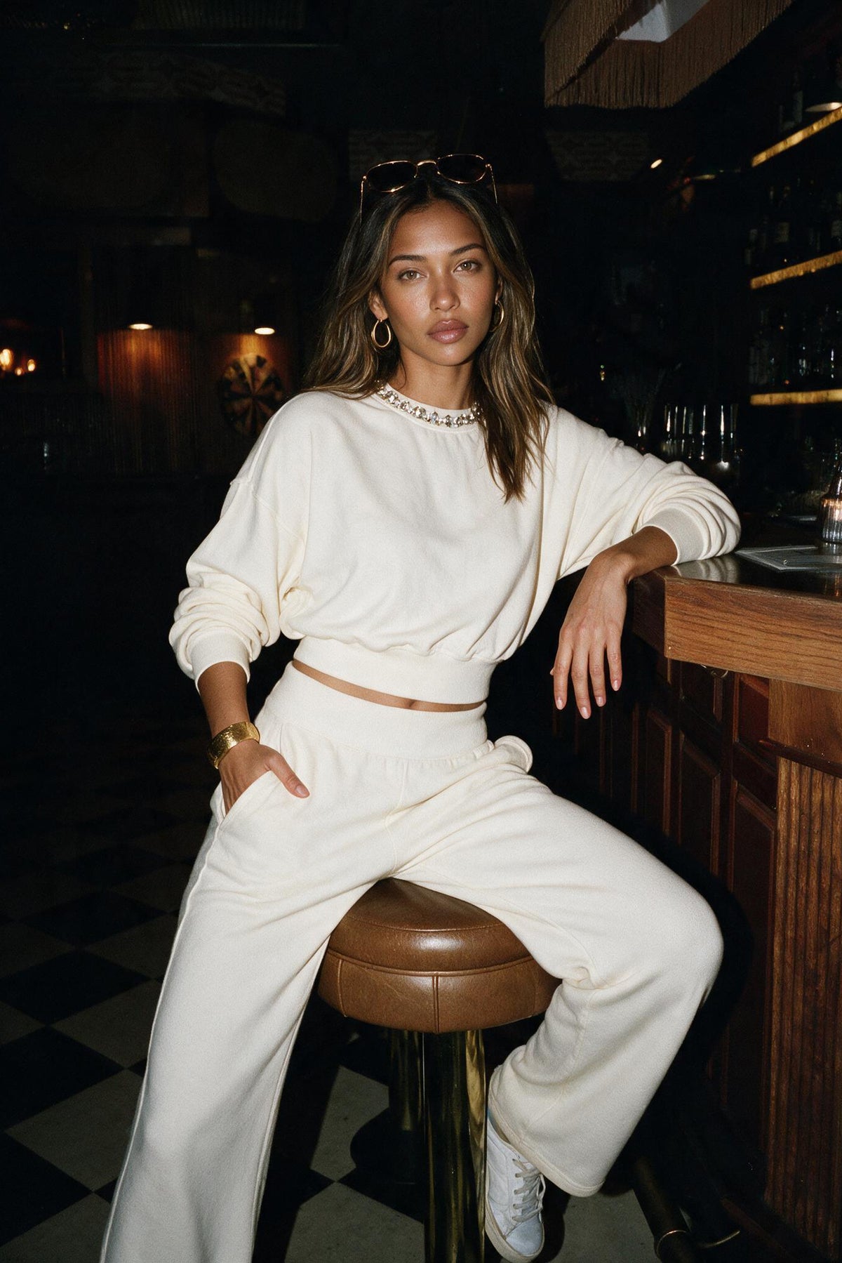 A woman wearing a matching cream-colored lounge set consisting of a long-sleeve cropped sweatshirt and wide-leg pants, sitting on a brown leather bar stool in a dimly lit bar environment.