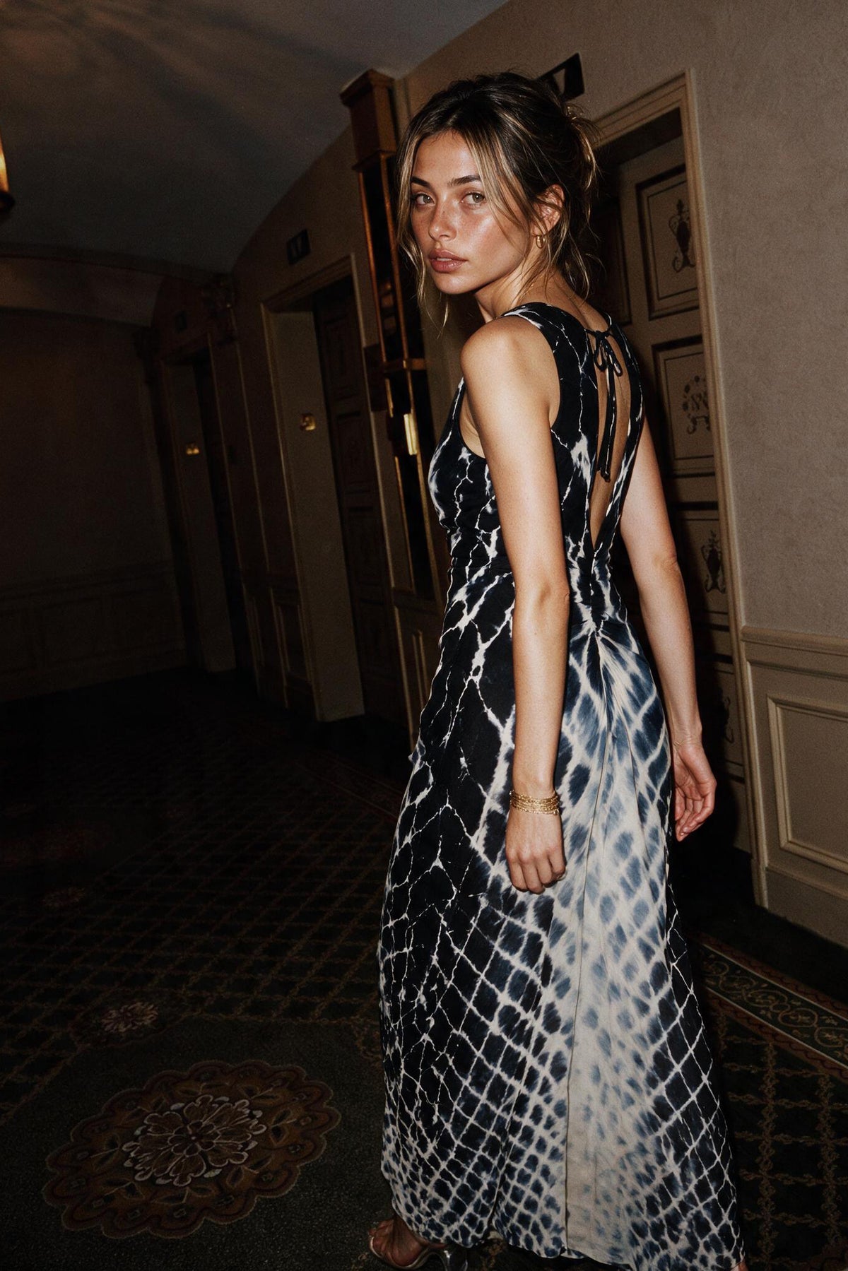 A woman wearing a sleeveless black and white patterned maxi dress with an open back, standing in a dimly lit indoor hallway.